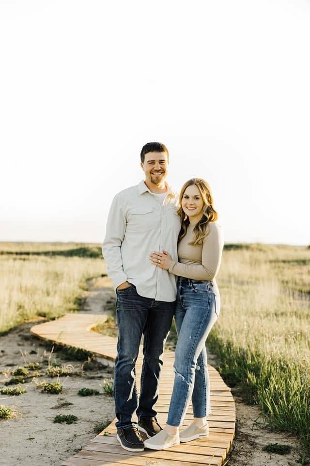 A smiling couple stands together on a wooden boardwalk in a vast, open grassland field at golden hour